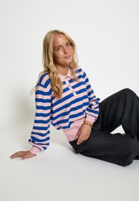 Pruhovaný svetr s vodorovnými modrými a růžovými pruhy, tříknoflíkovým zapínáním a žebrovanými manžetami. V kombinaci s volnými černými kalhotami.