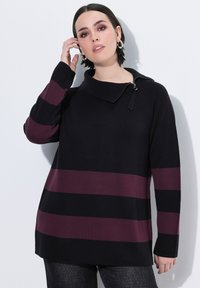 Kvinde med kort mørkt hår, der rører ved sit øre, iført sølv hoops øreringe og en sort sweater med maroon vandrette striber og en lynlås krave.