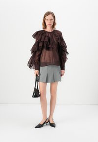 Transparante bordeaux blouse met ruches, gecombineerd met een geplooide grijze rok. Model houdt een kleine zwarte handtas vast en draagt zwarte puntige schoenen.