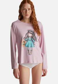 Joven mujer con cabello rojo largo lleva una camiseta de manga larga rosa que presenta a una niña de dibujos animados sosteniendo una linterna y una paloma blanca.