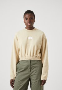 Felpa beige cropped con maniche lunghe, collarino a coste e orlo elasticizzato. Presenta una piccola etichetta con logo al centro. Indossata con pantaloni verdi.