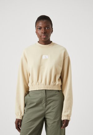 Beige cropped sweatshirt med lange ærmer, ribbet halsudskæring og elastisk nederste kant. Har et lille logo patch i midten. Bæres med grønne bukser.