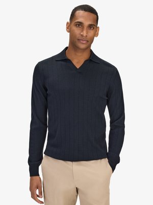 Man met navyblauwe geribbelde lange mouwen polotrui en beige broek, staand met handen gedeeltelijk in de zakken tegen een witte achtergrond.