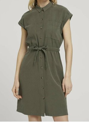 Femme portant une robe boutonnée vert olive à manches courtes, avec col, poche avant et ceinture nouée à la taille, debout devant un fond uni.