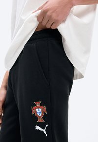 Κοντινό πλάνο ατόμου που σηκώνει λευκό πουκάμισο, αποκαλύπτοντας μαύρο παντελόνι με το έμβλημα της ποδοσφαιρικής ομάδας της Πορτογαλίας και λευκό λογότυπο Puma στο πάνω μέρος του μηρού.