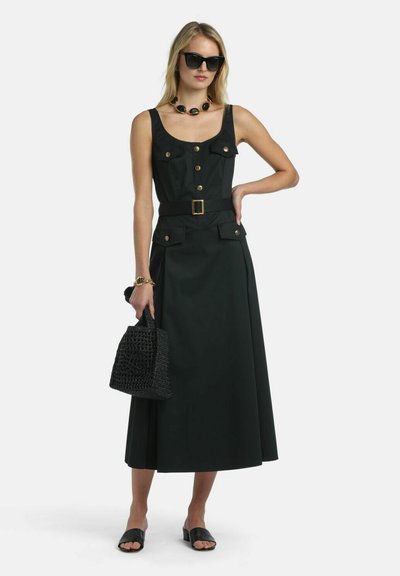 Frau trägt ärmelloses schwarzes Kleid mit goldenen Knöpfen und Gürtel, schwarze Sonnenbrille, hält eine schwarze gewebte Handtasche und trägt schwarze Sandalen.