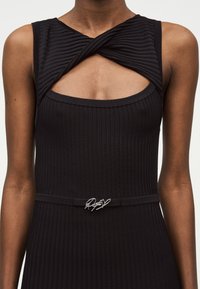 Close-up af en person iført en ærmeløs sort ribstrikket kjole med en snoet halsudskæring og et tyndt bælte med en sølv dekorativ spænde.