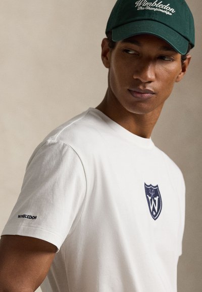 Άνδρας που φοράει λευκό T-shirt με λογότυπο του Γουίμπλεντον στο στήθος και το μανίκι, και σκούφο σε σκούρο πράσινο χρώμα με το λογότυπο του Γουίμπλεντον, κοιτάζοντας προς την πλευρά.