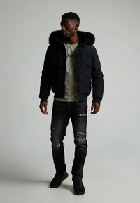 Zwarte pufferjas met een met bont gevoerde capuchon, doorgestikt ontwerp, gedragen over een lichtgrijs shirt, gecombineerd met versleten zwarte spijkerbroek en zwarte sneakers.