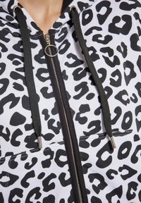 Witte en zwarte hoodie met luipaardprint, voorzien van een zilveren ritssluiting, zwarte trekkoorden en een voorzak voor extra functionaliteit.