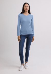 Himmelblaues Langarmshirt mit Rundhalsausschnitt, kombiniert mit dunklen Skinny-Jeans und weißen Sneakers, vor einem neutralen Hintergrund.