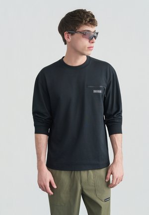 Tânăr care poartă o cămașă neagră cu mâneci lungi și buzunar cu fermoar pe piept și pantaloni verde măsliniu, uitându-se spre dreapta pe un fundal simplu.