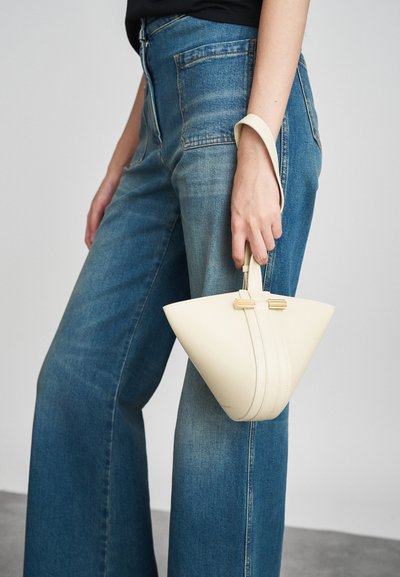 Konusa formas balta ādas somiņa ar rokturi, izceļoties ar redzamām šuvju detaļām un zelta apdari, apvienota ar augstiem ziliem džinsiem.
