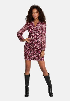 Femme aux cheveux bouclés portant une mini-robe à manches longues rose avec un motif et des bottes noires hautes jusqu'aux genoux, debout avec une main sur la hanche.