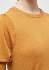 Detail zblízka na osobu v hořčicově žlutém pleteném topu s nafouknutými krátkými rukávy a žebrovaným kulatým výstřihem.