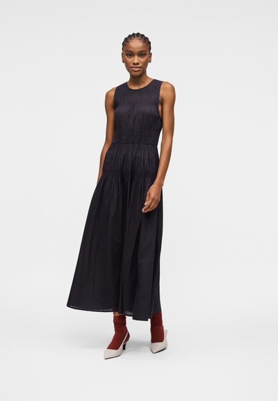 Femme aux cheveux tressés portant une robe midi noire sans manches, des chaussettes couleur rouille et des talons blancs, debout devant un fond uni.