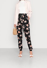 Joggings noirs à fleurs avec des fleurs orange et blanches, taille élastique, associés à un haut blanc et un blazer rose clair, tenant un sac tressé.