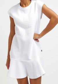 Vit kortärmad klänning med en utsvängd nederkant, prydd med en präglad Adidas-logotyp och en slät, mjuk tygkonstruktion.