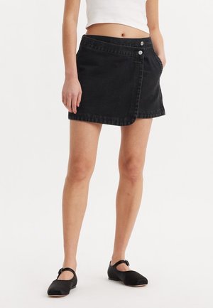 Femme portant une mini-jupe asymétrique noire et des ballerines noires Mary Jane, une main dans la poche de la jupe, sur fond blanc.