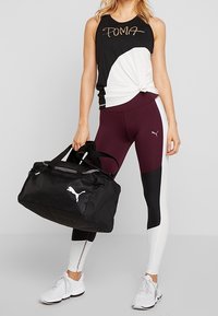 Top sin mangas de deporte en blanco y negro con detalle de nudo, mallas burdeos y negras, y una bolsa de deporte negra con logo blanco, combinado con zapatillas blancas.