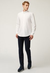 Camicia bianca a maniche lunghe con colletto, abbinata a pantaloni blu scuro e scarpe da ginnastica nere. Tessuto liscio, design sartoriale.