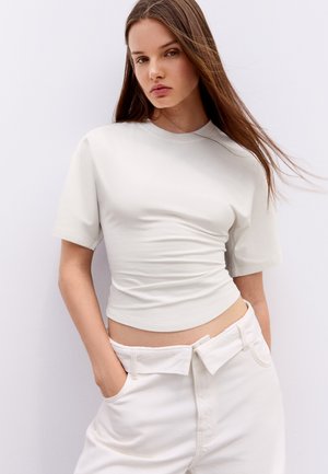 Jeune femme aux longs cheveux bruns portant un haut blanc ajusté à manches courtes et un pantalon blanc taille haute, les mains dans les poches.