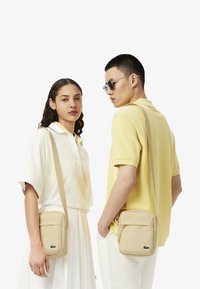 Beige Umhängetasche mit glatter Textur, ausgestattet mit einer vorderen Reißverschlusstasche und dem Lacoste-Logo. Tragen von Models in lässiger Kleidung.