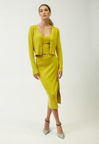 Strickensemble in leuchtendem Gelb mit einem figurbetonten Crop-Top, einem knöpfbaren Cardigan und einem Midirock mit seitlichem Schlitz. Glitzernde silberne Absätze ergänzen das Outfit.