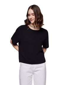 Top corto a maniche corte di colore nero in un tessuto leggero con una vestibilità comoda, caratterizzato da un collo rotondo e una texture sottile. Abbinato a pantaloni bianchi.