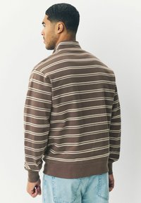 Brunstribet sweatshirt med krave, der har cremefarvede vandrette striber. Blød tekstur med ribbede mansjetter og linning. Lyseblå jeans synlige.