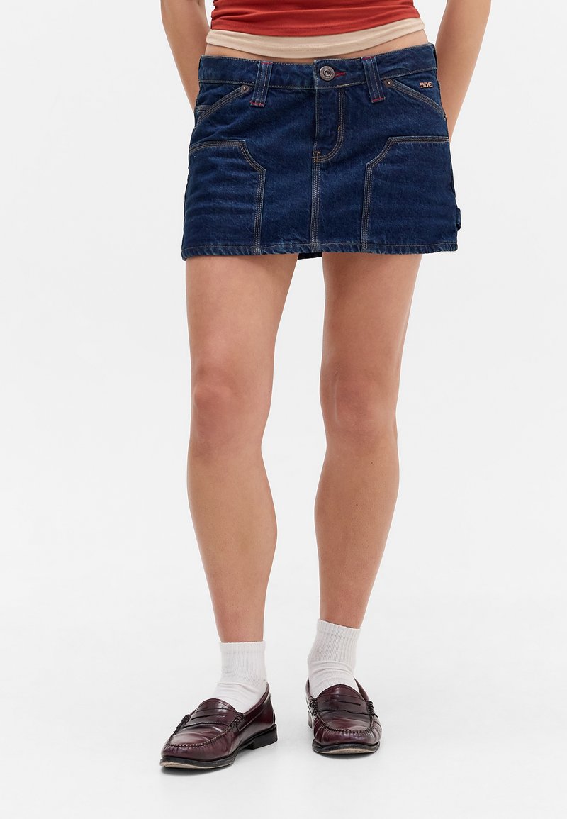 Denim krilo v temno modri barvi, ki vključuje sprednja žepa, zapiranje na gumb in kontrastno šivanje. Obuto z rjavimi loaferji in belimi nogavicami.
