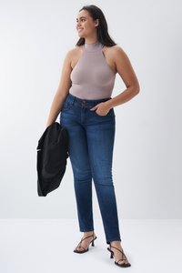 Bodysuit sin mangas de color taupe claro con cuello alto, combinado con jeans ajustados azul y sandalias de tacón negro con tiras. Sostenido de una chaqueta negra.
