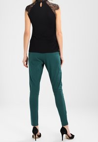 Top negro sin mangas con detalles de encaje en los hombros, combinado con pantalones verdes esmeralda y tacones altos negros. Textura suave, corte moderno.