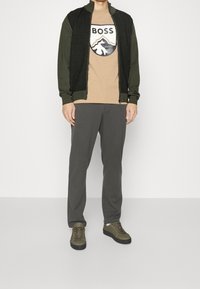 Oliivinvihreä vetoketjullinen takki, jossa on teksturoidut olkapäät, beige t-paita logolla, harmaat housut ja oliivinvihreät lenkkarit mustilla pohjilla.