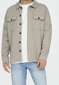 Light grey button-up shirt jacket made of soft fabric with a spread collar, two chest pockets, and black buttons. Worn over a white shirt.