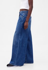 Modré široké džíny z denimu s vysokým pasem, výraznými švy, pěti kapsami a mírně třepeným lemem.