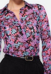Blusa de manga larga y botones, con un tejido negro translúcido decorado con vibrantes patrones florales en rosa, púrpura y azul. Incluye cuello y puños.