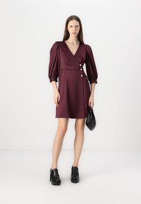 Mauve haljina omot s nabranim rukavima i tri dekorativna gumba; uparen s crnom torbicom i debelim crnim platformskim cipelama.
