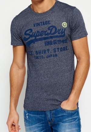 Mand iført en meleret navyblå SuperDry t-shirt og blå slidte jeans med den ene hånd i lommen mod hvid baggrund.