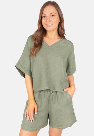 Femme portant un haut ample en lin vert olive à col en V et un short assorti avec cordon, debout, une main dans la poche, souriante.