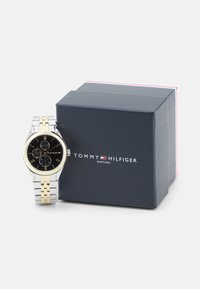 Montre en métal argenté et doré avec un cadran noir, trois sous-cadrans, index en chiffres romains et une boîte de marque en bleu foncé.