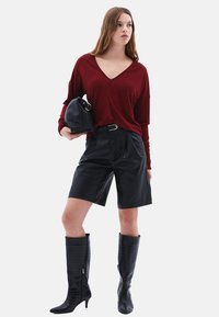 Maglione a V Bordeaux, pantaloni corti neri in pelle, stivali neri a ginocchio con effetto coccodrillo, mentre tiene una borsa nera, su uno sfondo bianco.