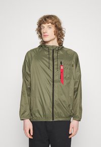 Olive green lightweight jacket with a hood, zippered front, elastic cuffs, and a chest pocket featuring a red tag. Smooth, shiny texture.