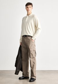 Μπεζ πόλο με μακριά μανίκια, καφέ cargo παντελόνι με πλάγιες τσέπες, μαύρη ζώνη και μαύρα παπούτσια. Μοντέλο που κρατάει μια σκούρα καφέ δερμάτινη τζάκετ.