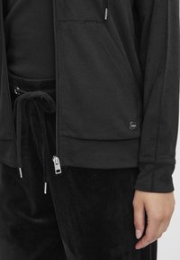 Veste zippée noire en tissu doux avec poches latérales, assortie à un pantalon noir avec taille élastique et texture lisse.