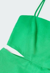 Top corto de lino verde con tirantes finos, que presenta un diseño sencillo y una textura suave. Nota el detalle de corte en el dobladillo.