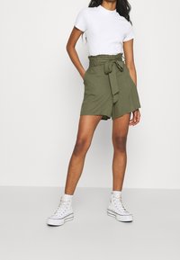 Olive green shorts with a high waist, belt tie, and pockets, paired with a white short-sleeved t-shirt and white high-top sneakers.