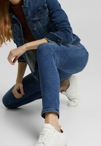 Chaqueta vaquera en azul medio, con un corte clásico, botones delanteros y bolsillos en el pecho. Jeans ajustados azules y zapatillas blancas completan el conjunto.