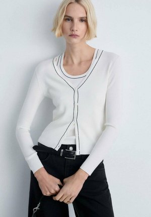 Cardigan bianco a coste con bordi neri, caratterizzato da una scollatura a V e chiusura con bottoni, abbinato a jeans neri e una cintura nera.