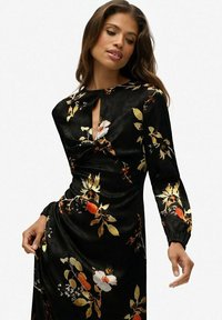 Czarna sukienka w kwiaty z intensywnymi pomarańczowymi, białymi i złotymi wzorami, z dekoltem w kształcie dziurki i długimi rękawami, o miękkiej, jedwabistej fakturze.